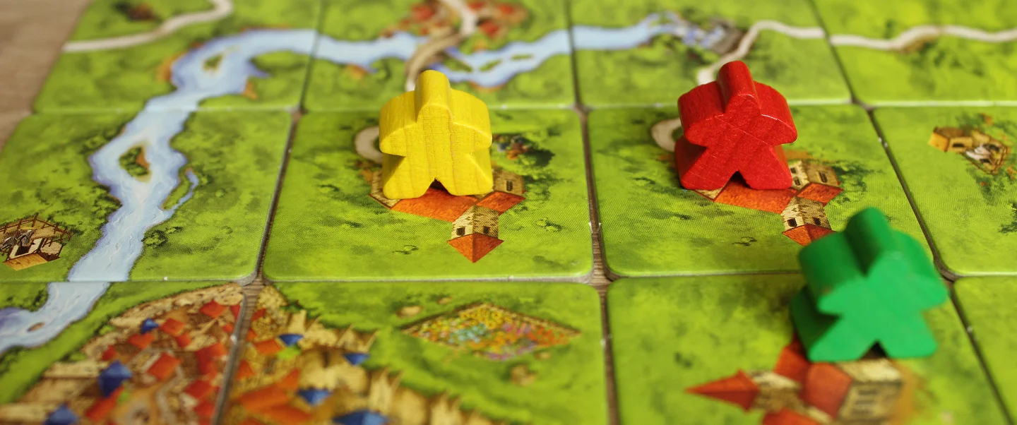 Una fotografia ravvicinata di un colorato gioco da tavolo in corso, con pedine di legno posizionate su tessere illustrate raffiguranti paesaggi con fiumi, villaggi e campi. Il vivace tabellone e la disposizione strategica dei pezzi evidenziano temi di pianificazione, competizione e gioco sociale da tavolo in un ambiente interno rilassato.